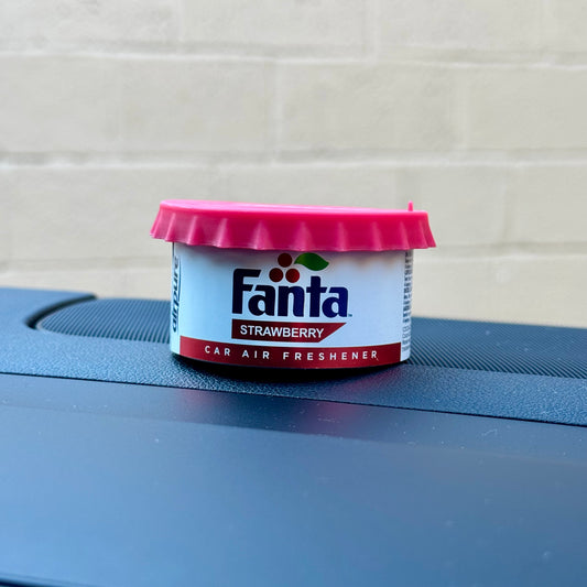 Fanta Strawberry Car Air Freshener Tin with twist-top lid, displayed on a car dashboard against a beige brick wall background.