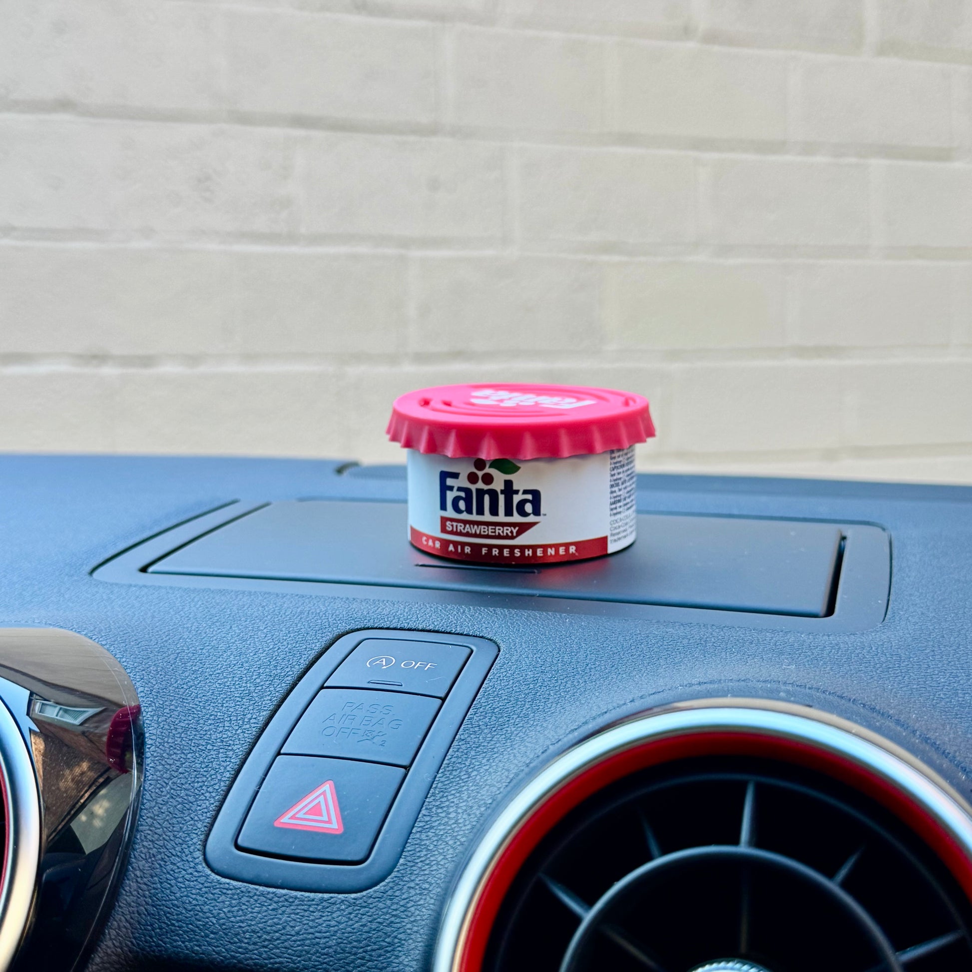 Fanta Strawberry Car Air Freshener Tin with twist-top lid, displayed on a car dashboard against a beige brick wall background.