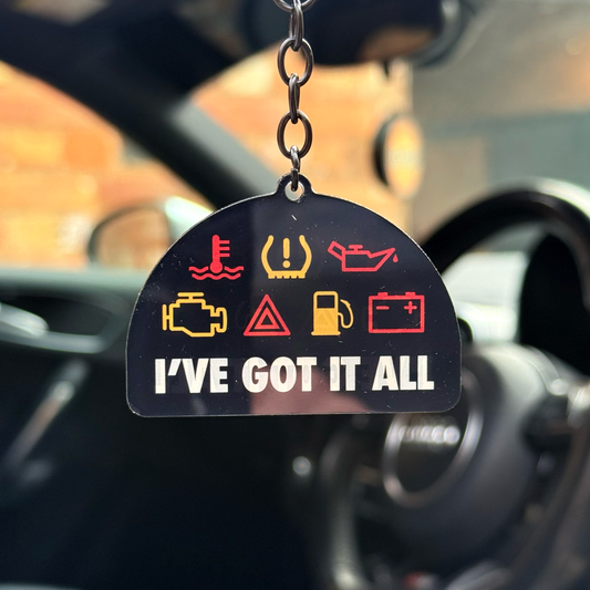Funny “I’ve Got It All” acrylic keychain hanging inside car with dashboard warning light symbols and gloss black chain