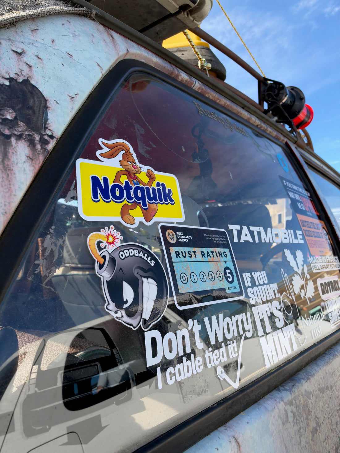 Funny vinyl stickers covering the side window of a rusty modified car, including Notquik and Rust Rating designs.