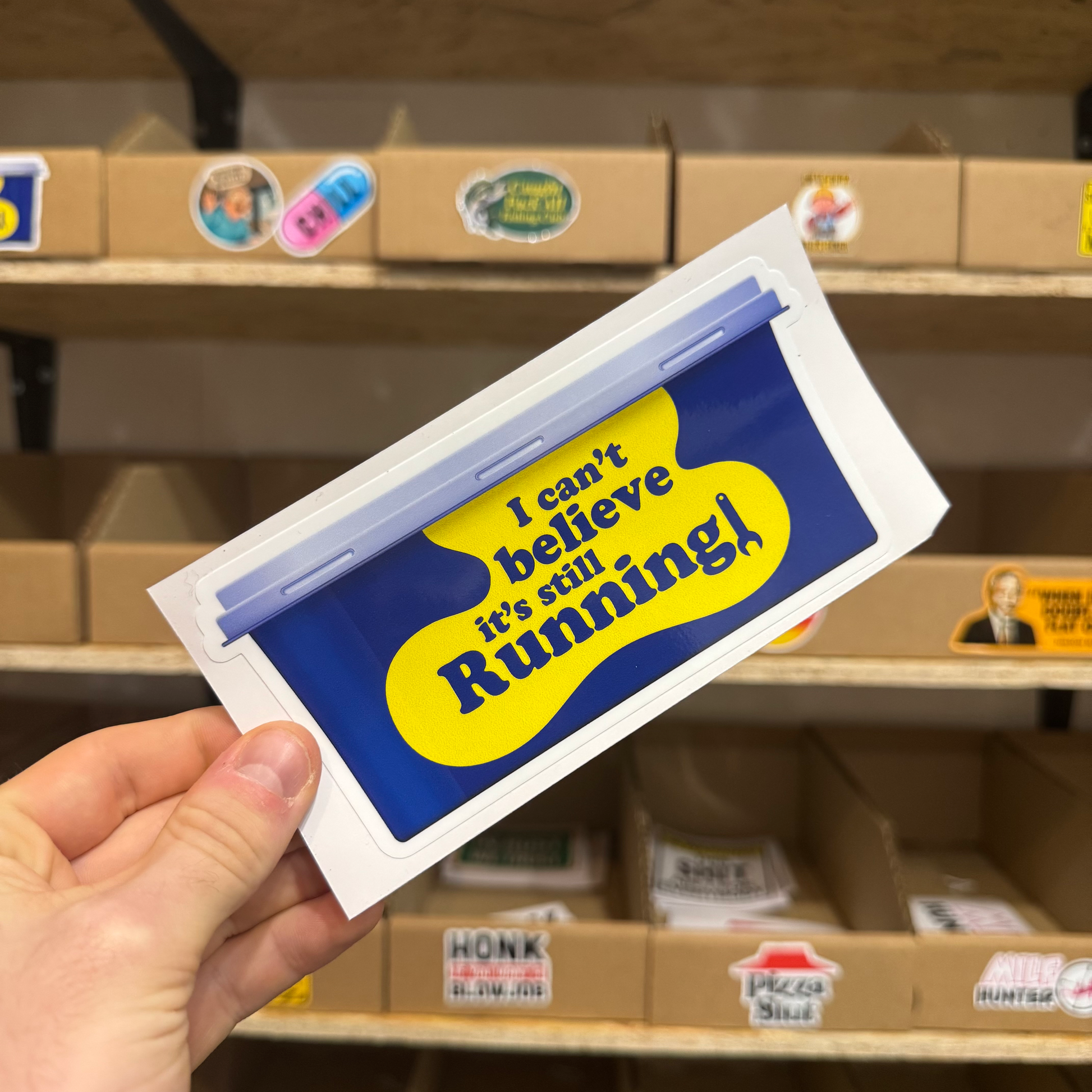 Blue and yellow parody car sticker with the text ‘I can’t believe it’s still running!’ designed to resemble a broken badge.