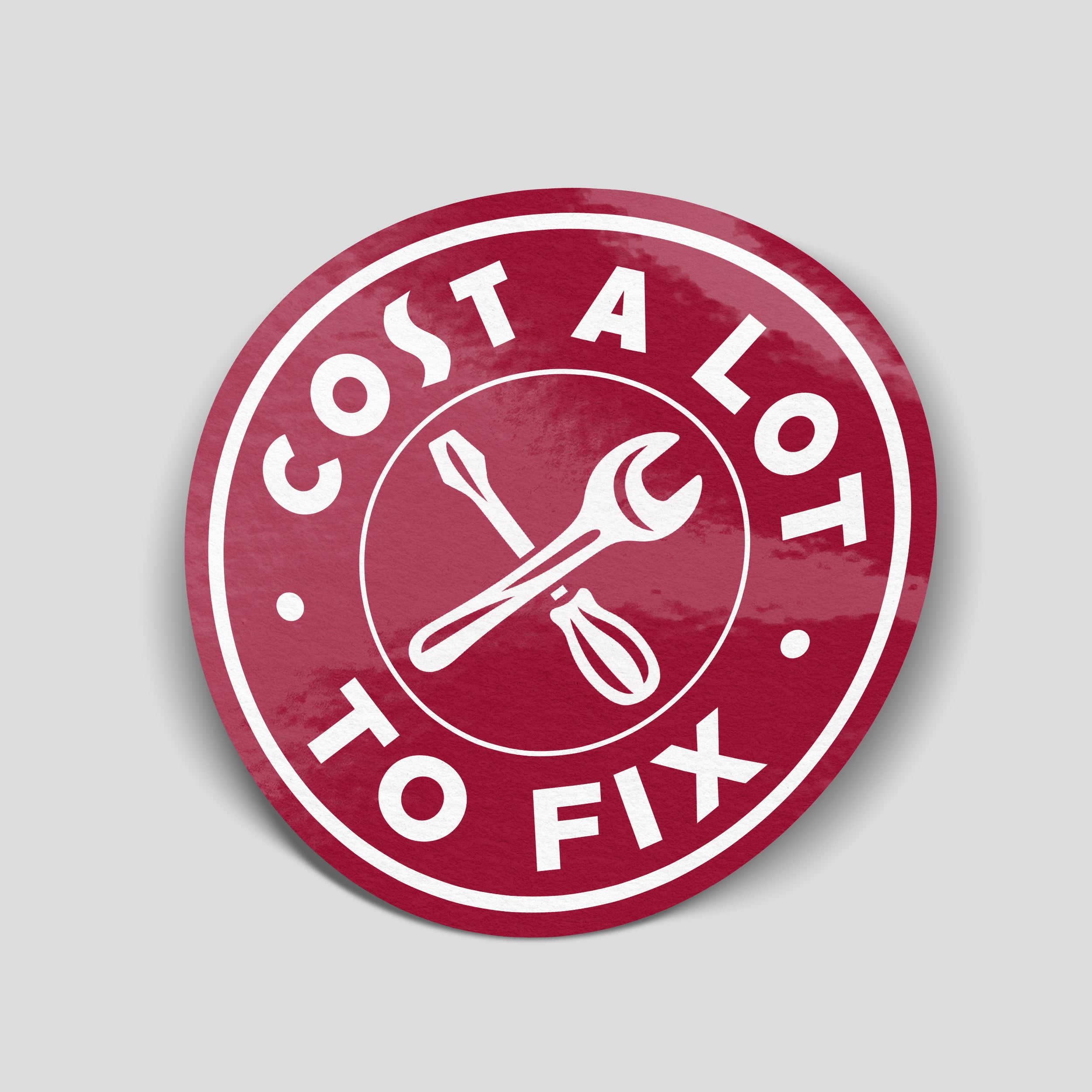 Burgundy circular “Cost A Lot To Fix” car sticker with white lettering and a crossed spanner and screwdriver.
