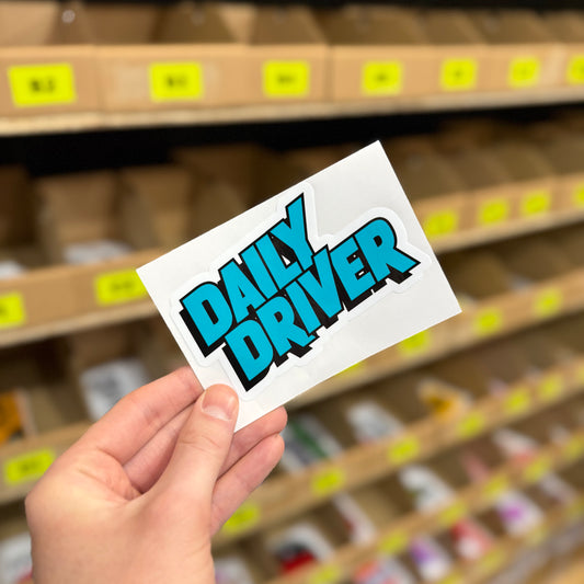 Daily Driver funny vinyl car sticker in bold turquoise and black text design, shown in hand against packing shelves. Car enthusiast decal printed on durable waterproof gloss laminated vinyl, designed and made in the UK.