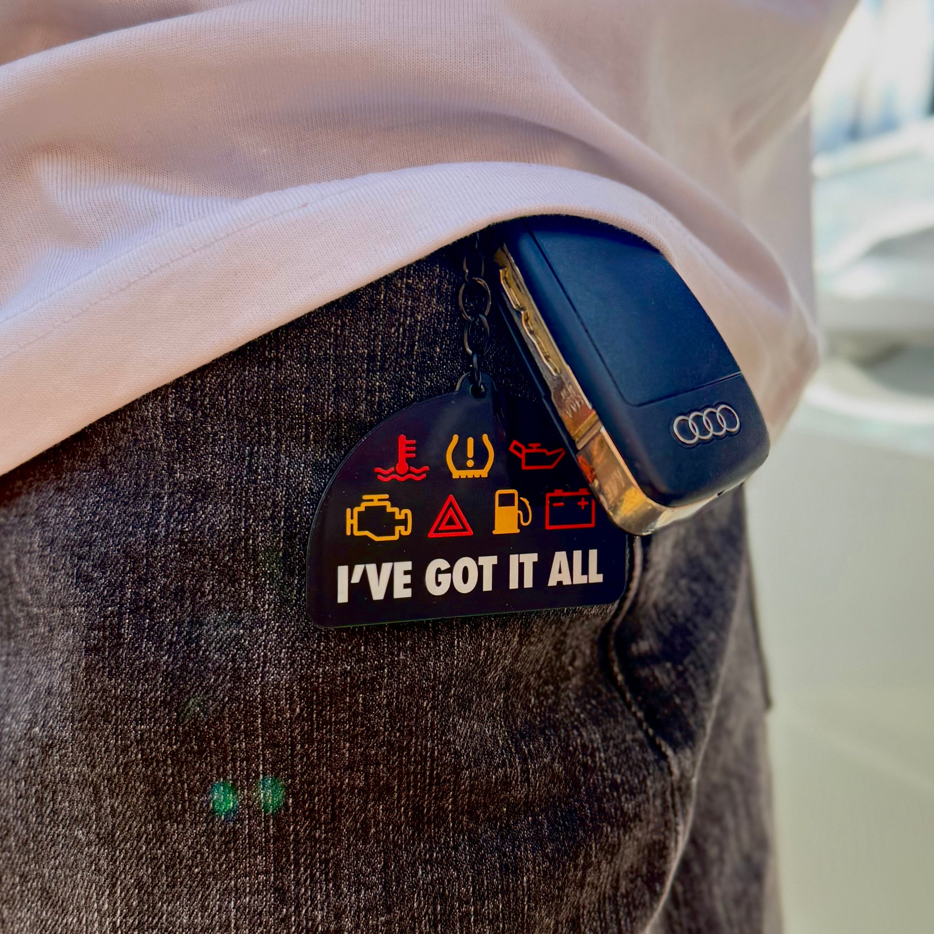 “I’ve Got It All” funny car warning light keychain attached to Audi car keys, shown hanging from a pocket beside a white car.