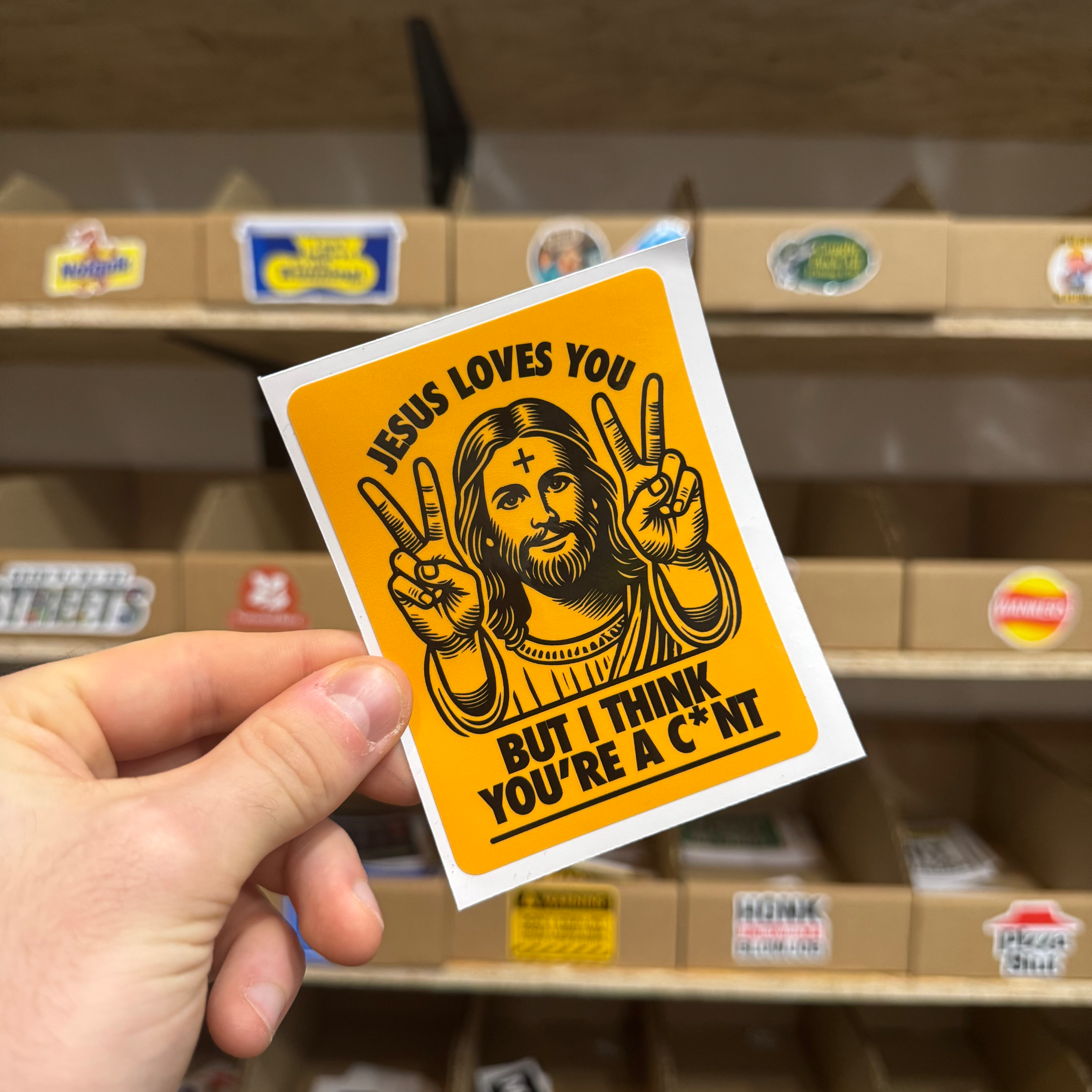 Bold orange sticker showing a cartoon Jesus holding up peace signs with the text ‘Jesus loves you but I think you’re a c*nt.’