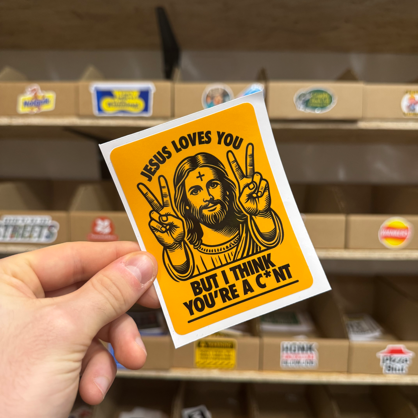 Bold orange sticker showing a cartoon Jesus holding up peace signs with the text ‘Jesus loves you but I think you’re a c*nt.’