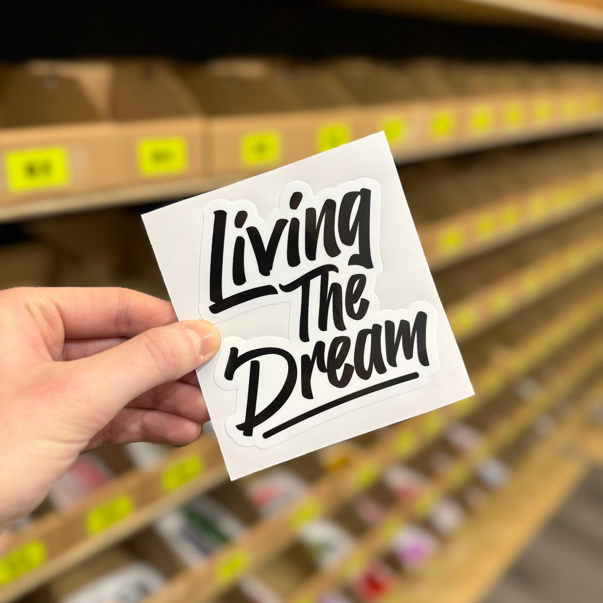 Black and white “Living the dream” text design funny vinyl sticker held in hand.