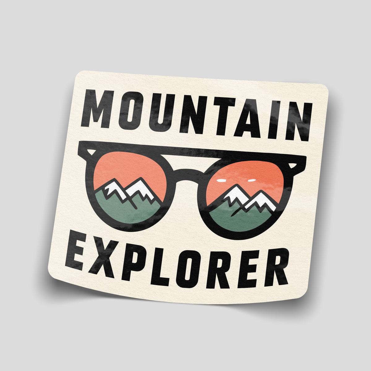 Cream Mountain Explorer sticker with black sunglasses showing green snow-capped mountains against orange skies in the lenses.