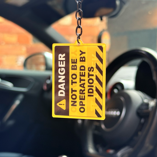 Yellow “Not To Be Operated By Idiots” funny acrylic keychain hanging in car with gloss black chain and keyring
