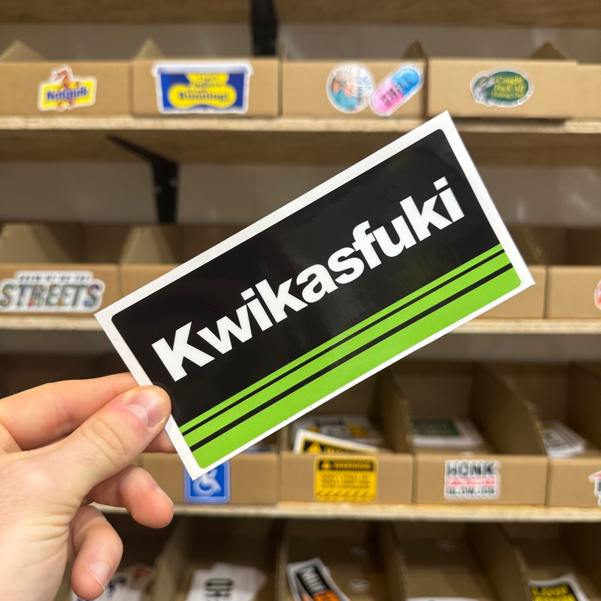 Motorbike parody sticker with a black background, white text ‘Kwikasfuki,’ and green racing stripes.