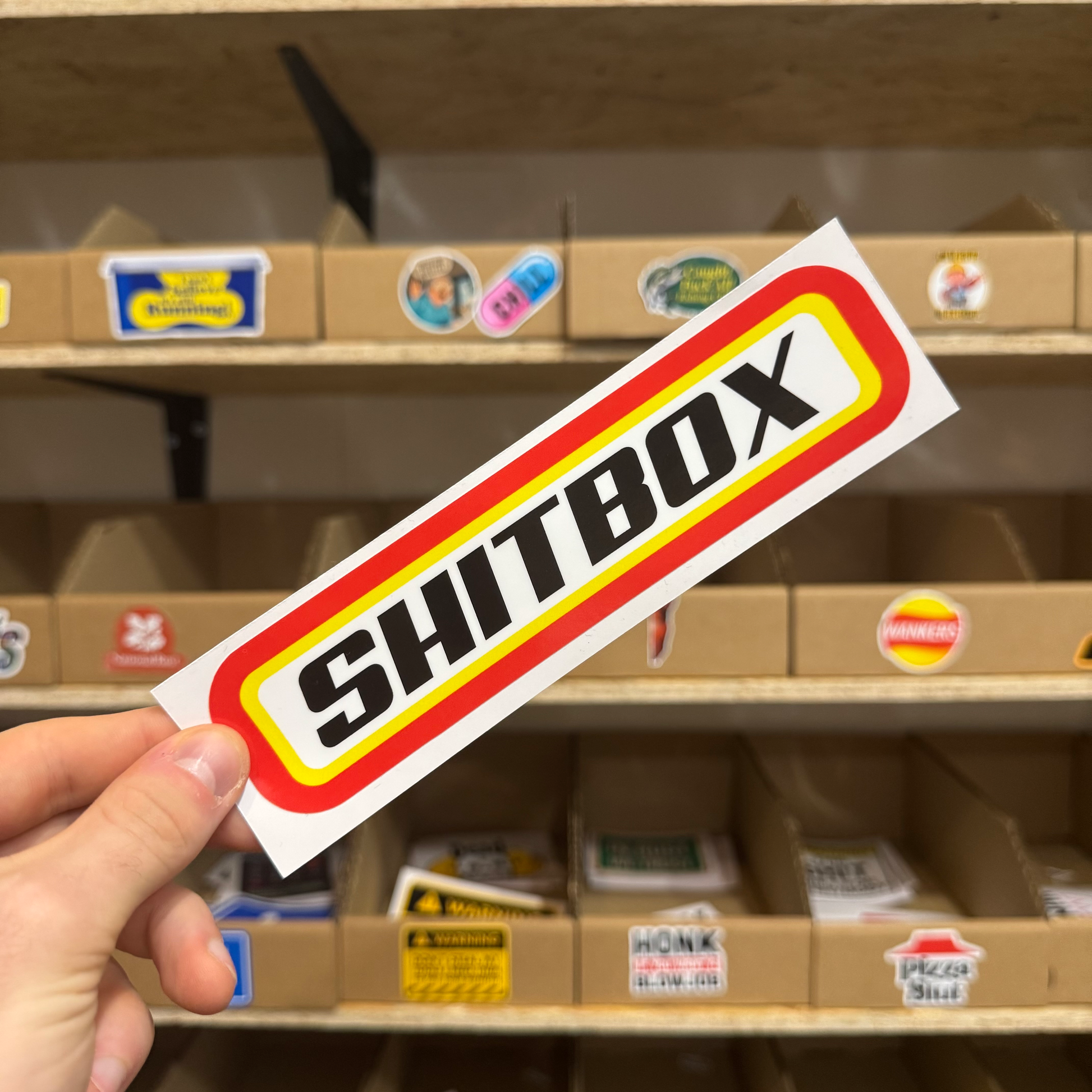 Retro-style yellow and red sticker reading ‘Shitbox,’ ideal for old or beat-up cars.