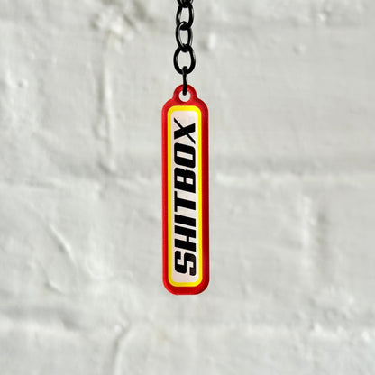 Funny “Shitbox” acrylic keychain with bold red and yellow design, glossy double-sided print and black chain, perfect for project car owners.