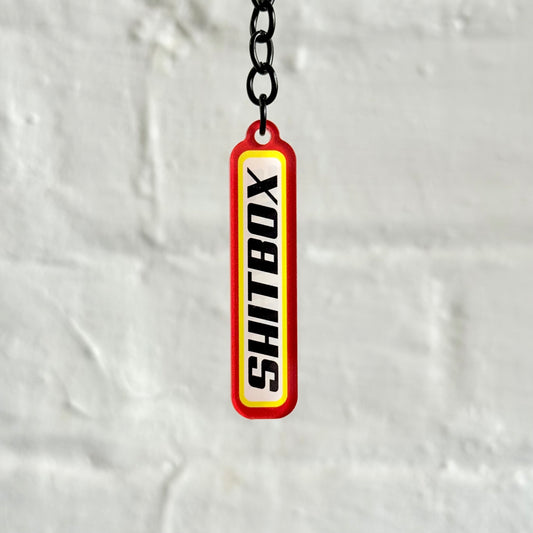 Funny “Shitbox” acrylic keychain with bold red and yellow design, glossy double-sided print and black chain, perfect for project car owners.