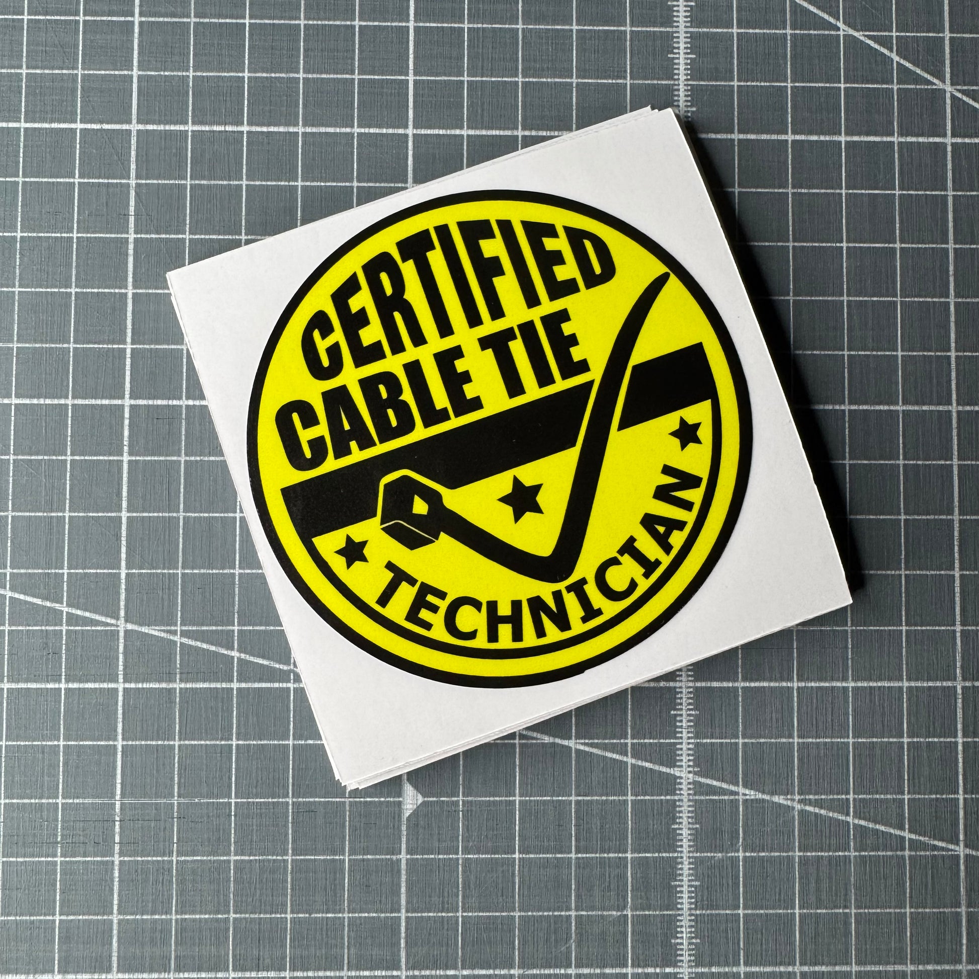 Stack of circularYellow and black certified cable tie technician vinyl decal stickers for cars, toolboxes or as a funny sticker joke. Featuring bold text and a cable tie in the shape as a tick.