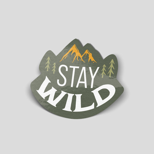 Olive green Stay Wild adventure sticker with large white lettering, golden mountain peaks and pale green pine trees.