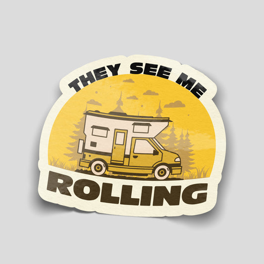 They See Me Rolling sticker featuring a yellow and cream motorhome, pine trees and bold dark lettering against a golden background.
